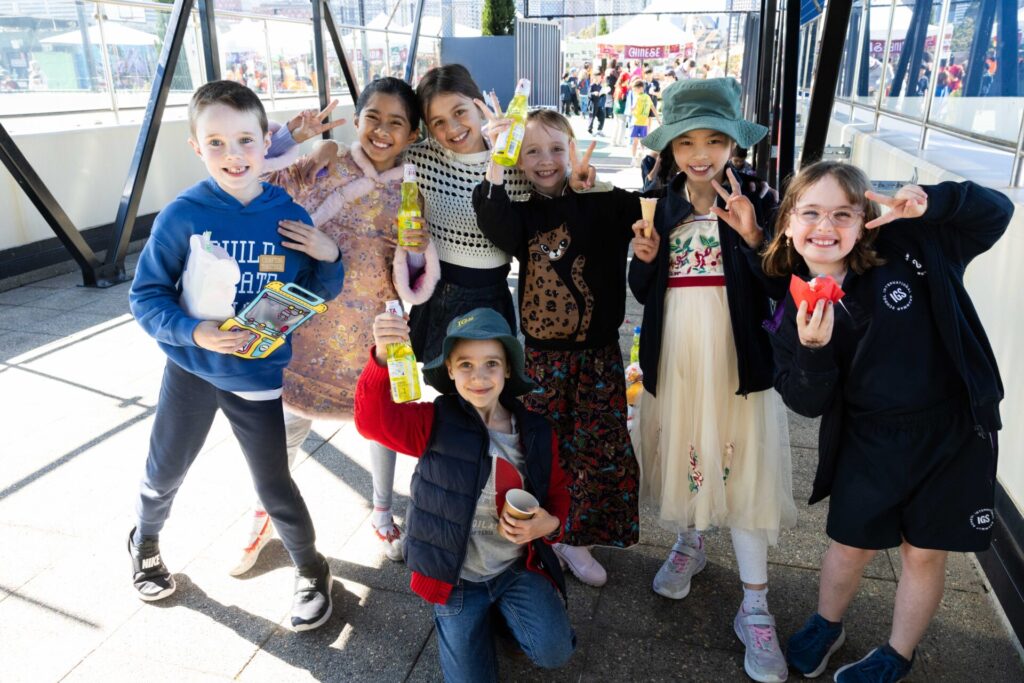 Seven smiling children pose together outdoors, holding drinks and snacks, some making peace signs. Casually dressed, they stand on a sunny day at a country festival with colourful tents and other people in the background—capturing the joy found in all stories of friendship and celebration.