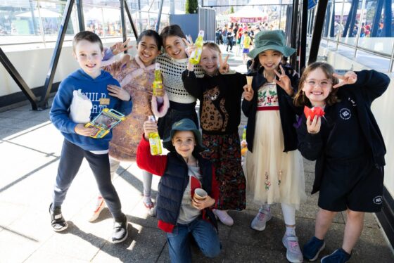 Seven smiling children pose together outdoors, holding drinks and snacks, some making peace signs. Casually dressed, they stand on a sunny day at a country festival with colourful tents and other people in the background—capturing the joy found in all stories of friendship and celebration.