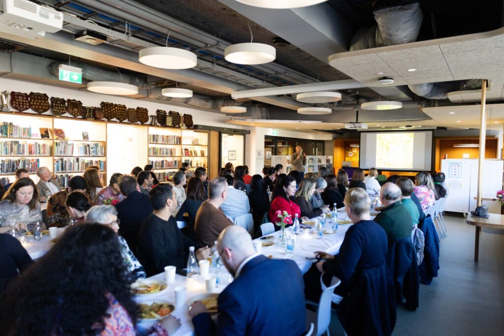 A large group of people sit at round tables in a library, listening to a speaker at the front as a presentation is projected onto a screen. Shelves of books and award plaques line the walls, celebrating achievements from across the country where all stories begin.