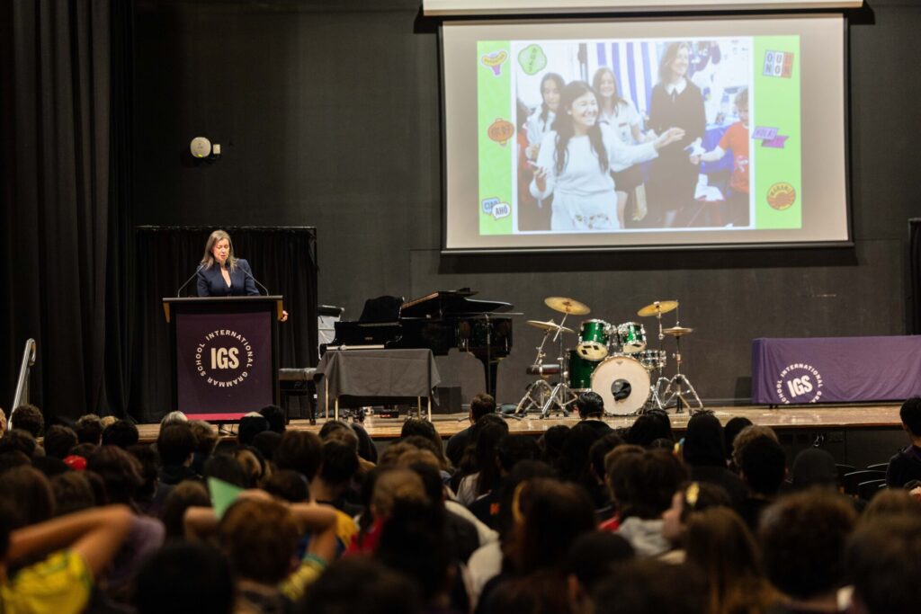 A woman speaks at a lectern labelled IGS on stage, with an audience watching. Behind her, a screen displays a group photo and musical instruments—including a drum kit and piano—setting the scene for the country to begin sharing all stories that unite its people.