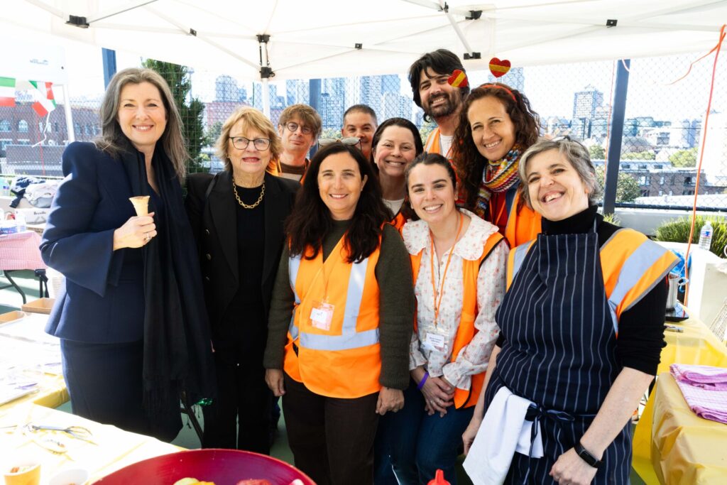 A group of people, some wearing orange safety vests, smile together under a white marquee at an outdoor event with city buildings in the background, capturing the spirit of Term in Full Flight.