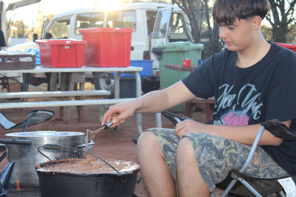 A young person in a black T-shirt stirs food in a large pot outdoors, sitting on a camping chair. Camping equipment and a white vehicle are visible in the background.