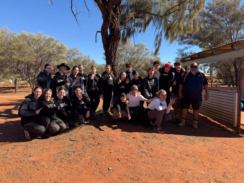 A group of people pose together outdoors on red sandy ground, surrounded by trees. Some are sitting or crouching at the front whilst others stand behind, all smiling at the camera under a sunny sky near a shelter.