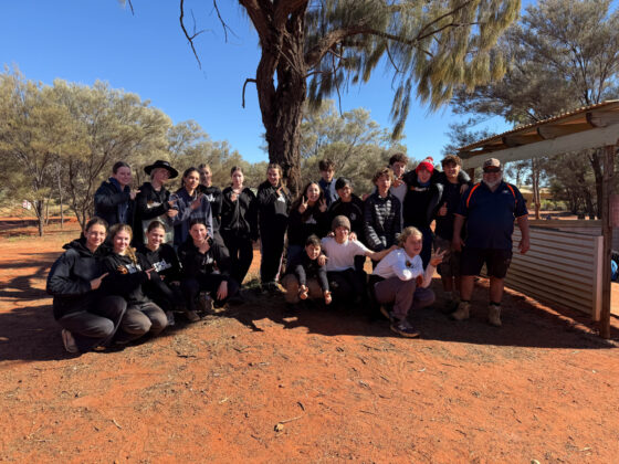 A group of people pose together outdoors on red sandy ground, surrounded by trees. Some are sitting or crouching at the front whilst others stand behind, all smiling at the camera under a sunny sky near a shelter.