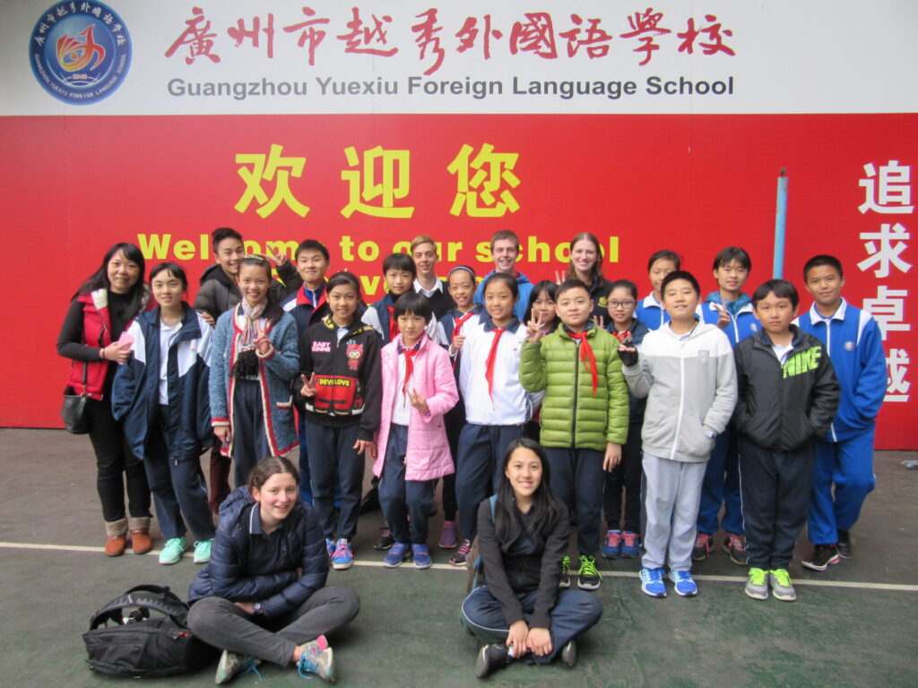 A group of students and adults, representing a vibrant global community, pose together outside in front of a red sign for Guangzhou Yuexiu Foreign Language School, smiling for the photo.