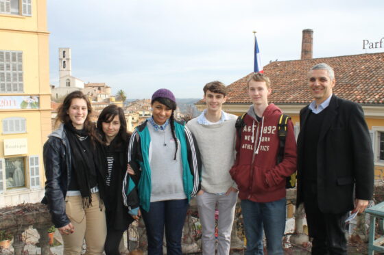 Six people stand close together outdoors, smiling at the camera. Behind them are buildings, rooftops, and a partially visible flag. The group appears relaxed and happy, embodying a sense of global community. The sky is slightly overcast.