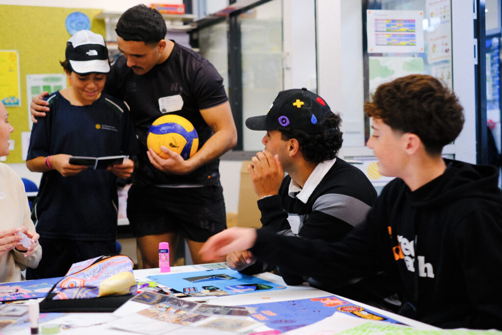 Four young people gather round a table covered with colourful papers and photos. One holds a volleyball and hugs another, who looks at a notebook. Two others sit and watch, smiling. Together, they share ideas in what appears to be a classroom, building health and creating healthier futures as they collaborate.