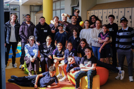 A group of children and adults, some in school uniforms, gather indoors in front of lockers, smiling and posing together on colourful furniture and standing in the background—a vibrant scene that highlights the spirit of health collaboration for building healthier futures.