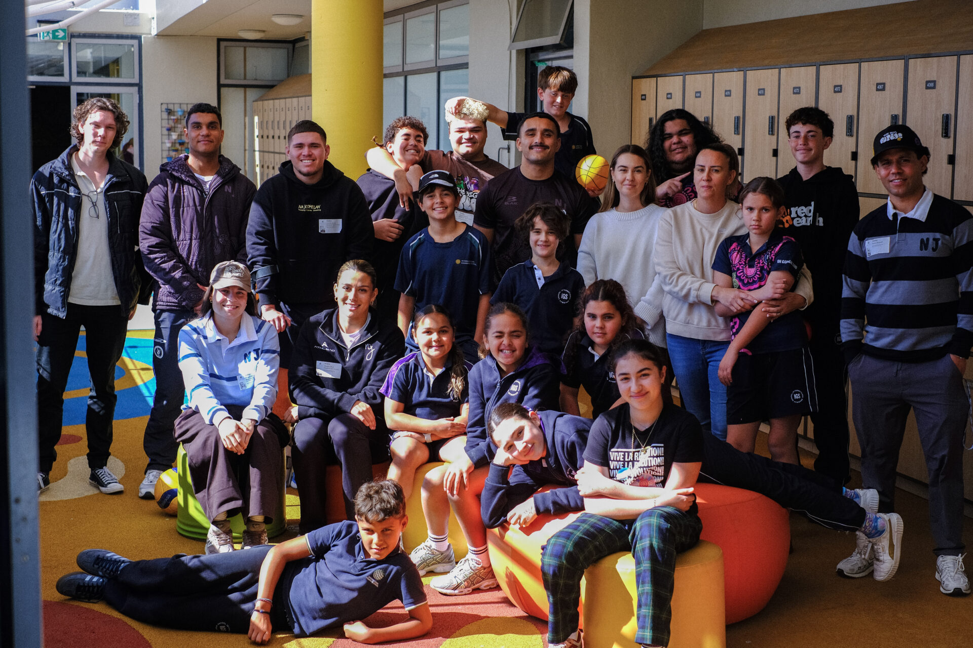 A group of children and adults, some in school uniforms, gather indoors in front of lockers, smiling and posing together on colourful furniture and standing in the background—a vibrant scene that highlights the spirit of health collaboration for building healthier futures.