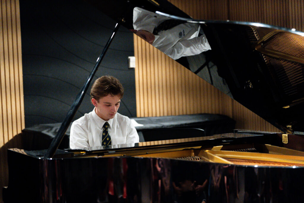 A young person in a white shirt and tie plays a grand piano in a modern room with wood panelling and black acoustic panels, creating a motivational sound that echoes the spirit of the Sound of Success.