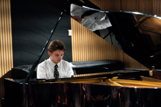A young person in a white shirt and tie plays a grand piano in a modern room with wood panelling and black acoustic panels, creating a motivational sound that echoes the spirit of the Sound of Success.