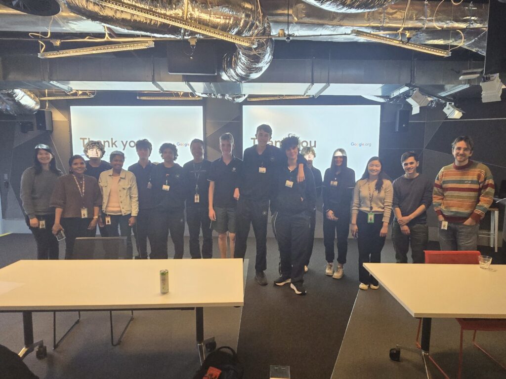 A group of people stand and smile for a photo in a modern office space with exposed ducts, embodying the collaborative spirit of Inside Google. Two screens behind them display Thank you. Tables and chairs are in the foreground.