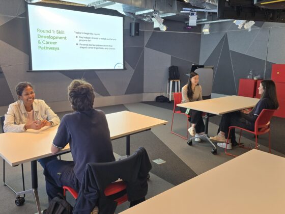 Four people sit in pairs at two tables having discussions in a modern, brightly lit room. A screen displays Round 1: Skill Development & Career Pathways along with discussion prompts, offering a glimpse Inside Google’s approach to collaborative learning and professional growth.