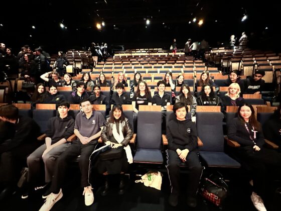 A group of students sits in several rows of blue theatre seats, posing for a photo. The background features more empty seats, with a few people standing and talking—capturing a lively moment before Stories on Stage begins.