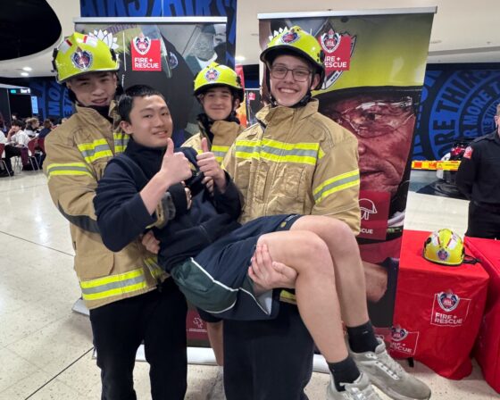 Four smiling teenagers pose at a fire and rescue event. Three wear firefighter gear and helmets, and together they lift and hold up a fourth friend, who gives a thumbs-up. Fire and rescue banners and table are visible in the background, creating an engaging setting for a powerful road safety lesson.