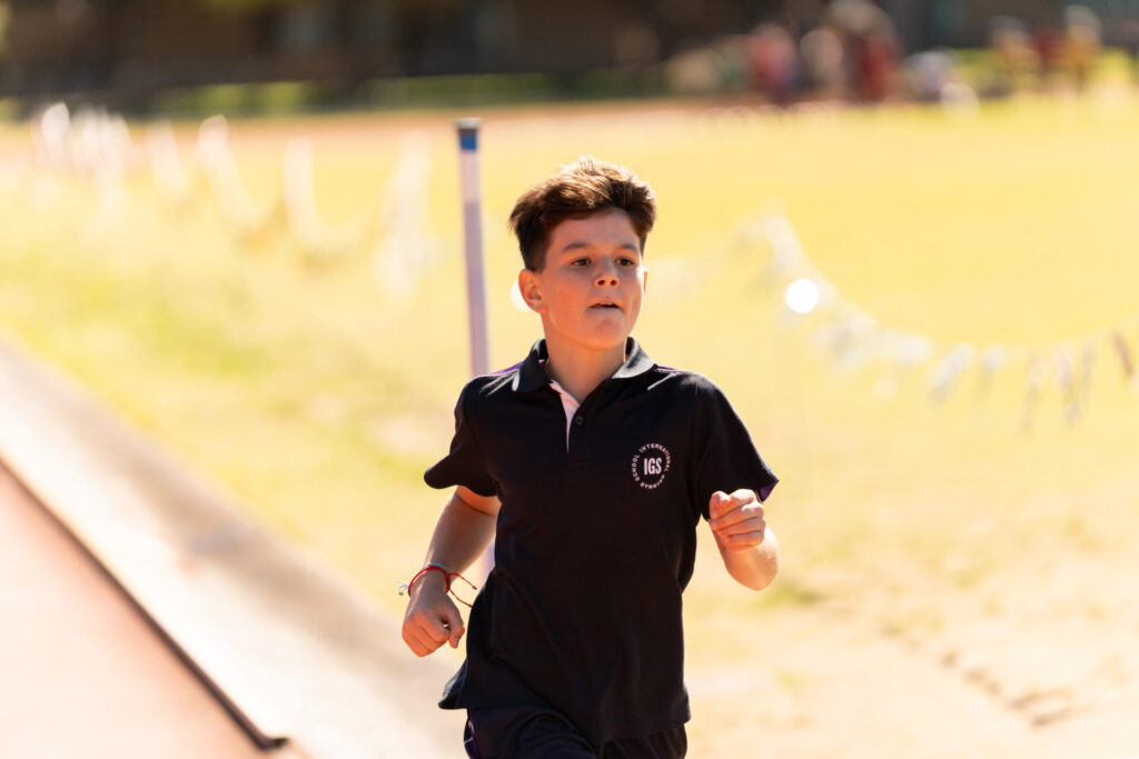 A young boy wearing a navy blue shirt runs on an outdoor track in bright sunlight, with a blurred background of grass and distant people.