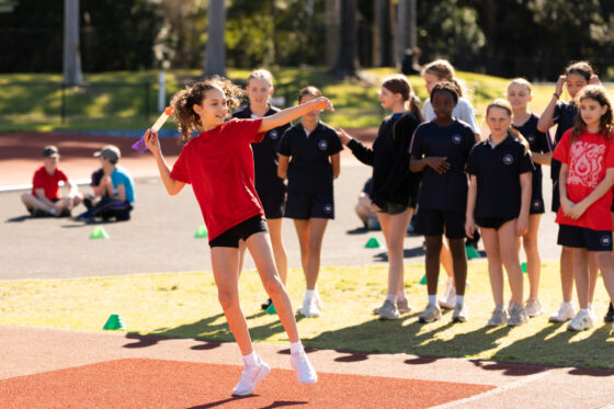 A girl in a red shirt prepares to throw a relay baton on a track, whilst a group of children in black uniforms watch in the background.