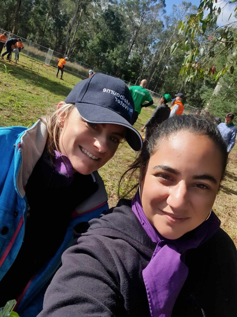 Two women wearing purple scarves pose for a selfie outdoors—one, possibly OZLEM HUSEYIN, wears a dark cap and blue jacket. People in orange vests work in the grassy, wooded background under a sunny sky, perhaps as part of the 2003 WITWAT event.