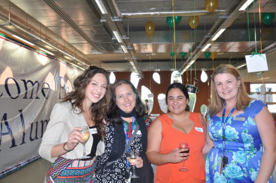 Four women stand close together, smiling and holding drinks at an indoor WITWAT event decorated with balloons and a large banner in the background, commemorating 2003 and featuring OZLEM HUSEYIN.