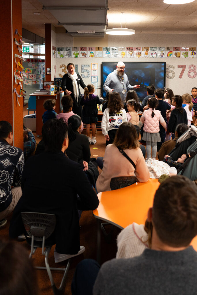 A group of children stand near a nursery teacher and a large screen, while parents watch from tables in a colourful classroom decorated with numbers and art, creating a warm welcome for everyone.