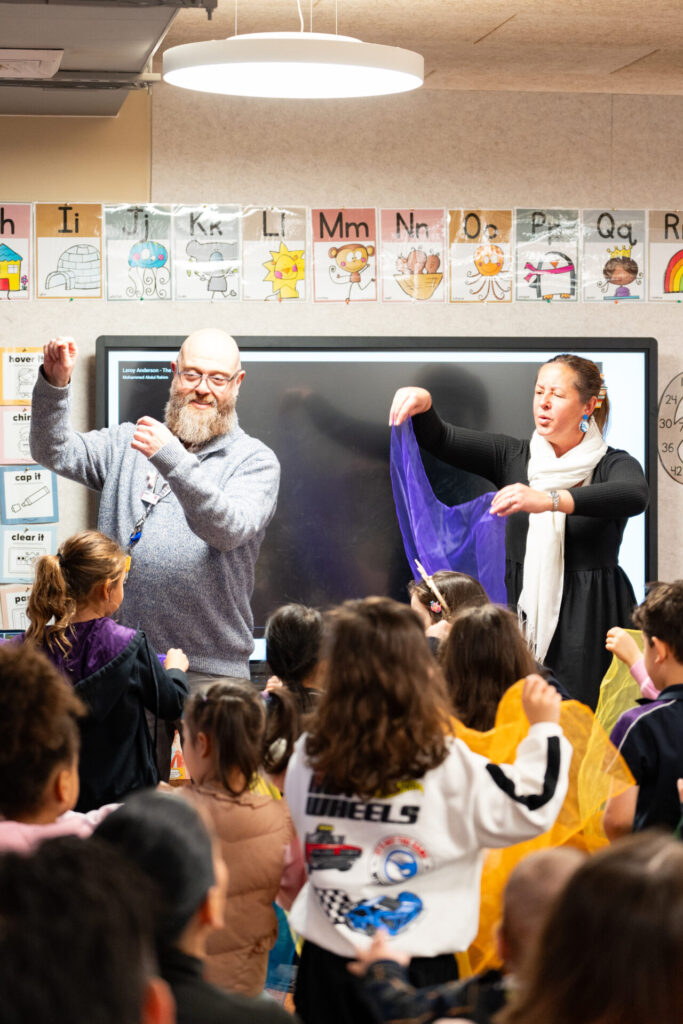A male teacher and a female teacher lead an engaging activity with colourful scarves in a kindergarten classroom full of young children. Alphabet cards and children’s artwork are displayed on the wall behind them, creating a warm welcome for everyone entering the room.