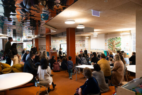 A group of adults and children sit in a colourful nursery classroom, facing a speaker at the front. The walls are decorated with educational posters and artwork, and the atmosphere appears lively and engaged, offering a warm welcome to all attendees.