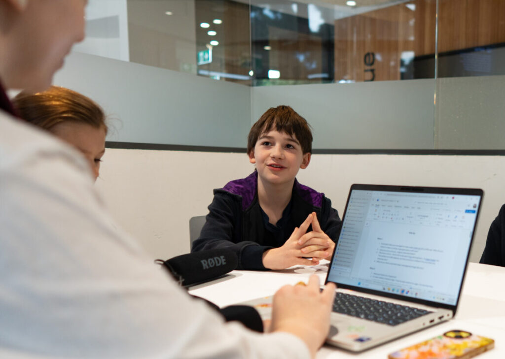 Four IGS students sit around a round table with a laptop, engaged in discussion or group work in a modern classroom or meeting space, collaborating on an assignment inspired by ABC Kids Listen.