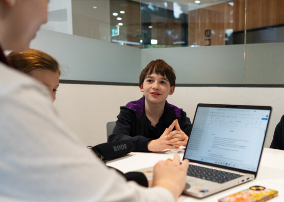 Four IGS students sit around a round table with a laptop, engaged in discussion or group work in a modern classroom or meeting space, collaborating on an assignment inspired by ABC Kids Listen.