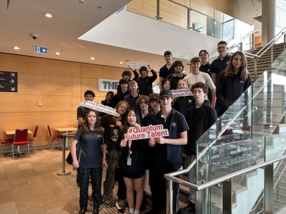 A group of young people pose on a staircase indoors, holding quantum-themed signs. They are smiling and appear to be part of a Quantum Futures programme or event related to quantum technology or education.