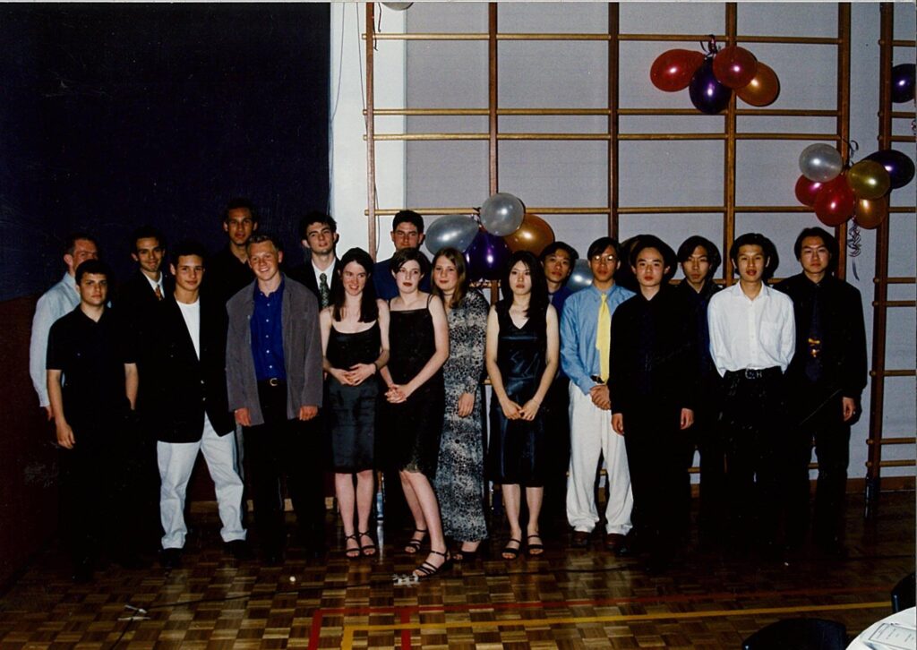 A group of young people dressed formally stand in a row indoors, with balloons and a wooden gym wall behind them, posing for a group photo at a celebration or dance.