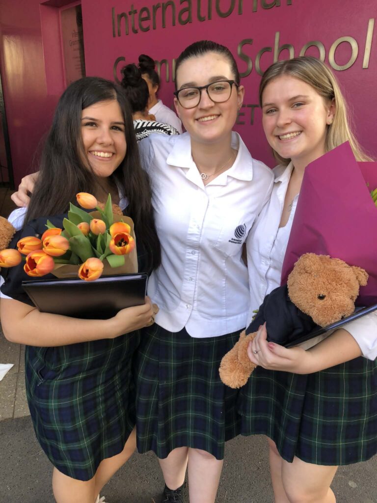Three young women in plaid skirts and white shirts stand smiling in front of a pink wall. Two hold bouquets and one holds a teddy bear, suggesting a celebratory event, possibly a graduation or school ceremony.