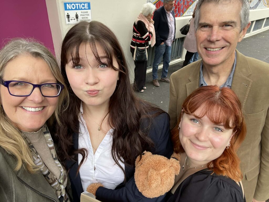 Four people smile for a selfie outdoors. One young woman pulls a playful face and holds a teddy bear. An IMPORTANT notice about hygiene is visible behind them. Other people stand in the background near a wall.