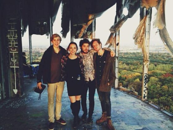 Four friends stand together, smiling, inside a damaged, graffitied building overlooking a forest and cityscape in the background. The roof above them is torn and ragged.