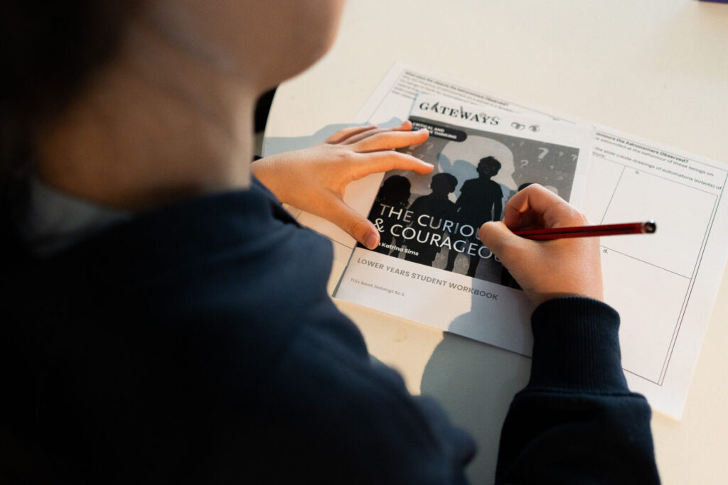 A student writes with a red pencil on a worksheet titled The Curious & Courageous: Lower Years Student Workbook at a light-coloured desk.
