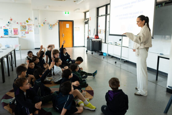A teacher stands at the front of a classroom, speaking to a group of seated young pupils, some of whom have their hands raised. Classroom decorations and a projector screen are visible in the background.
