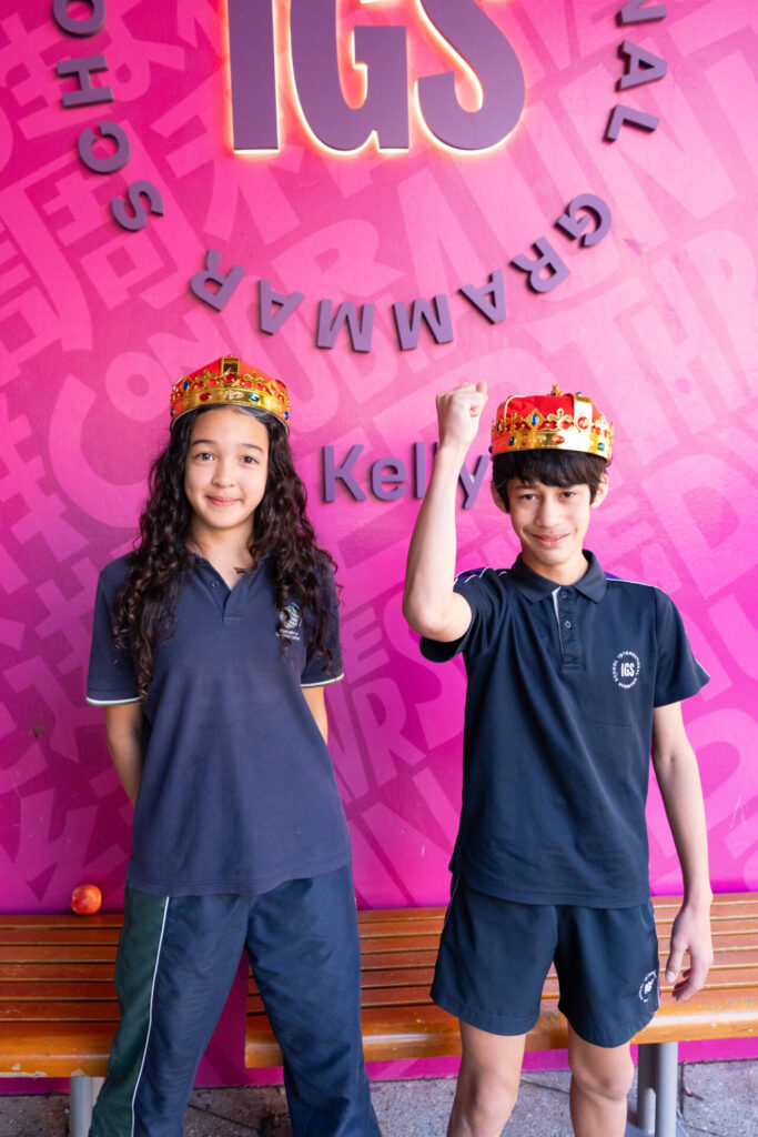 Two students in navy uniforms and crown props stand in front of a pink wall with IGS and other text. The student on the right raises a fist in triumph, while the other stands with hands by their sides. Both are smiling, seemingly basking in multilingual applause as they celebrate achievements recognised across five languages.