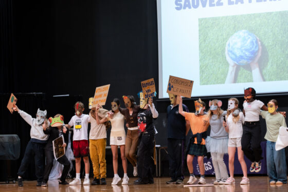 A group of children wearing animal masks hold protest signs and pose on stage in front of a screen showing an image of Earth and French text, as the audience erupts in multilingual applause.