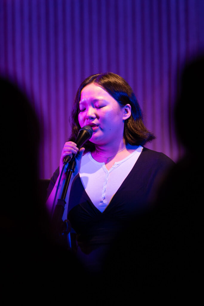 A person sings passionately into a microphone on stage, bathed in purple and blue lighting during the HICES Festival, with blurred silhouettes of an audience in the foreground.