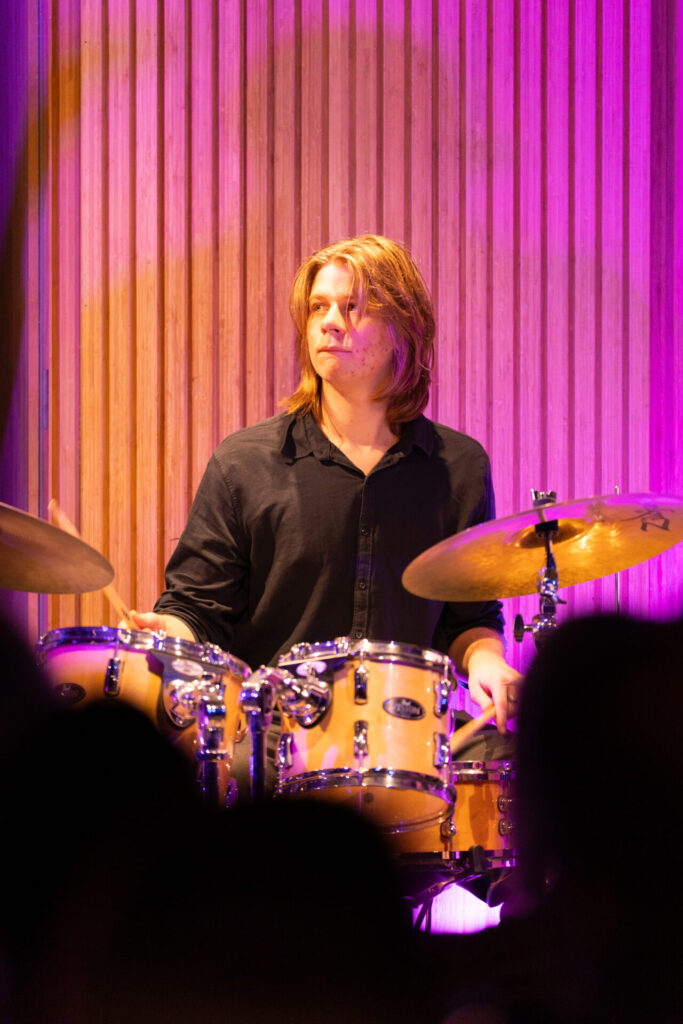 A drummer with long hair in a black shirt plays a drum kit on stage, immersed in purple lighting and set against a wooden panelled background, capturing the vibrant energy of the HICES Festival.