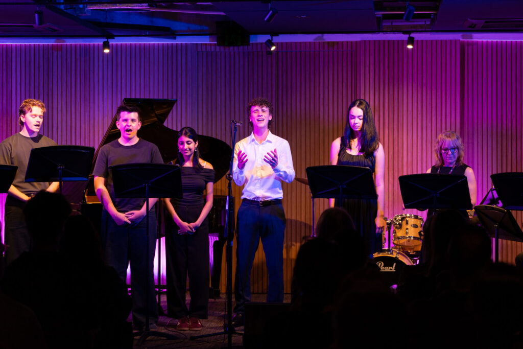 Six people stand on stage, each at a music stand, performing under purple lighting in this vibrant HSC Showcase. A piano and drums are visible behind them, highlighting the musical atmosphere. The audience is seated in the foreground, immersed in this exciting celebration of music.
