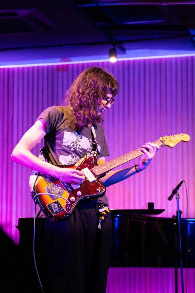 A person with long hair and glasses plays an electric guitar on stage under purple and pink lighting, with a microphone and piano in the background, capturing the vibrant energy of the HICES Festival Music HSC Showcase.