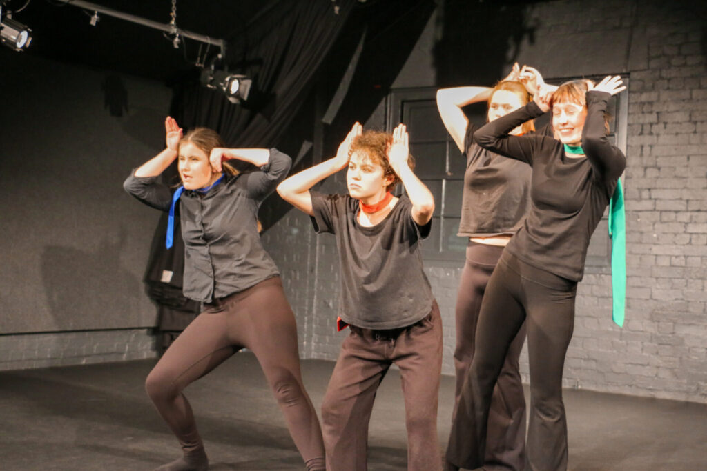 Four people on a stage, dressed in black with coloured scarves, hold their hands to their heads like animal ears, performing in front of a dark brick wall under stage lights—an expressive moment from their HSC Drama final act.