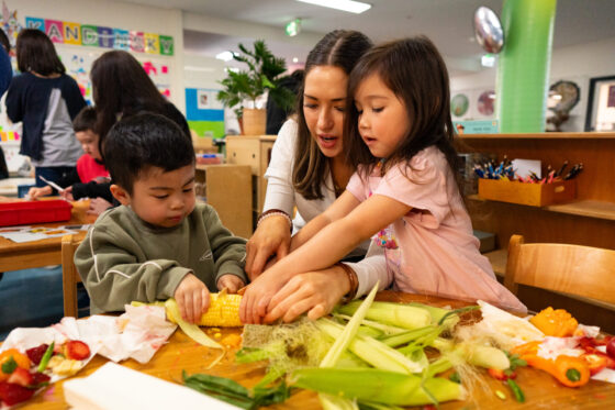 A teacher helps two young children peel sweetcorn at a table covered with vegetables in a bright, colourful classroom, nurturing their Seeds of Curiosity during Science Week at IGS.