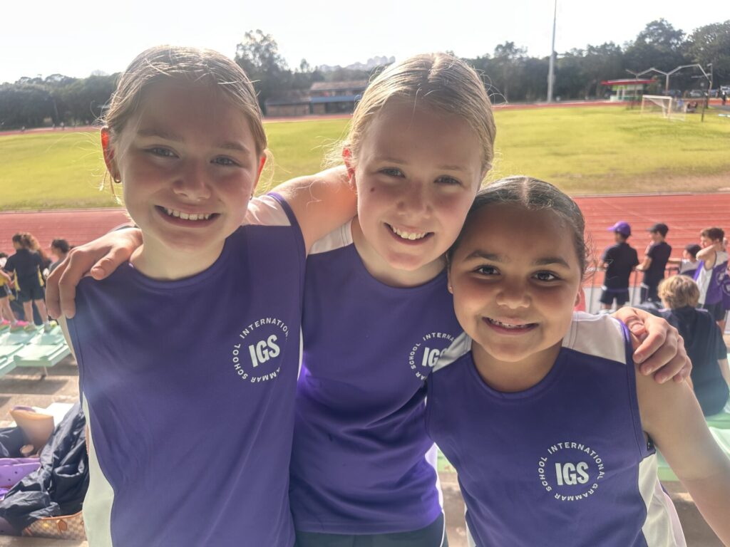Three smiling children in purple IGS uniforms stand together on terracing at an outdoor athletics track, arms around each other, with sunlight shining on them—truly on track for success. Other people and a green field are visible in the background.