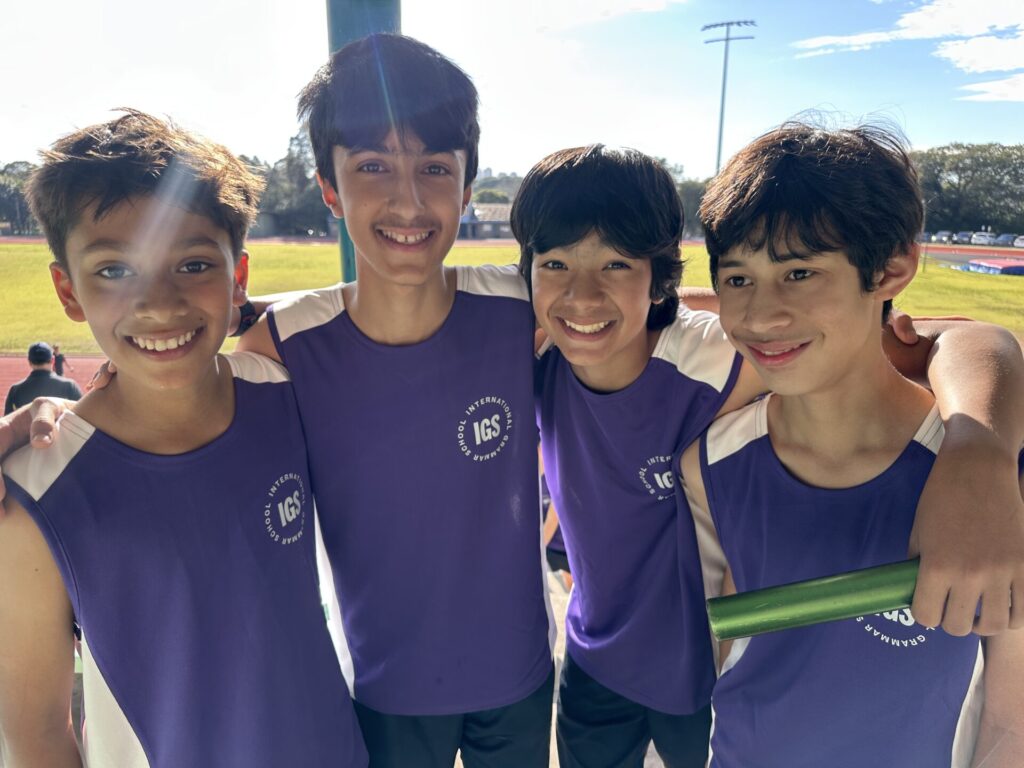 Four smiling boys in matching purple athletic shirts stand side by side, one holding a green relay baton, at an outdoor sports field on a sunny day, proudly showing they are on track for success.