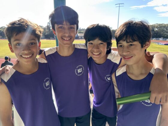 Four smiling boys in matching purple athletic shirts stand side by side, one holding a green relay baton, at an outdoor sports field on a sunny day, proudly showing they are on track for success.