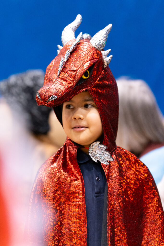 A young child wears a shiny red dragon costume with silver horns and green eyes, standing indoors with a blurred blue background and people nearby.
