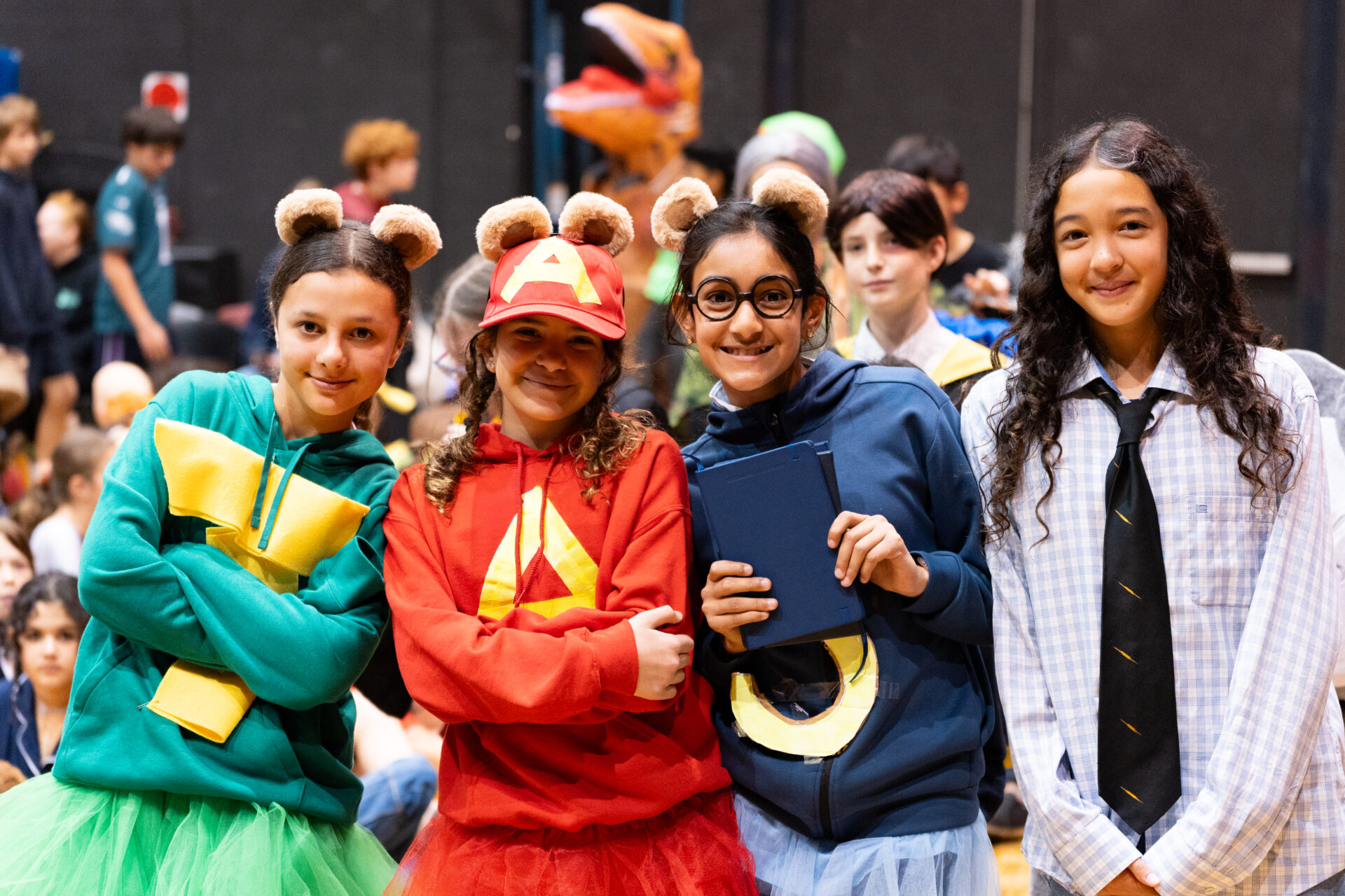 Four children in colourful bear-themed costumes pose and smile at an indoor event with a crowd in the background. Three wear bear ears and tutus, and one wears a long shirt and tie.