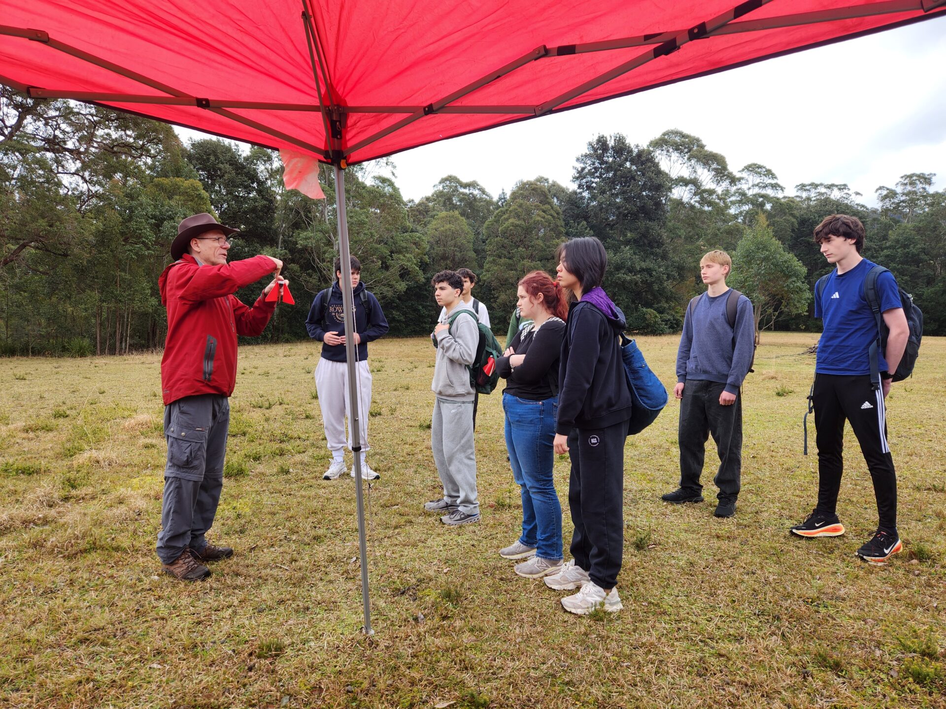 A man in a red jacket talks to a group of six teenagers standing under a red canopy in a grassy field, surrounded by trees, as he shares survival tips for the outdoors.