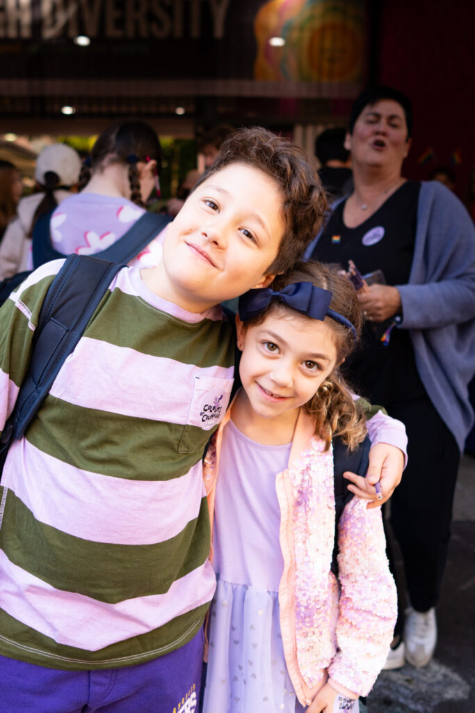 Two smiling children stand close together, one with an arm round the other. They are outside among a crowd, surrounded by the Colours of Courage reflected in the vibrant and lively backgrounds behind them.